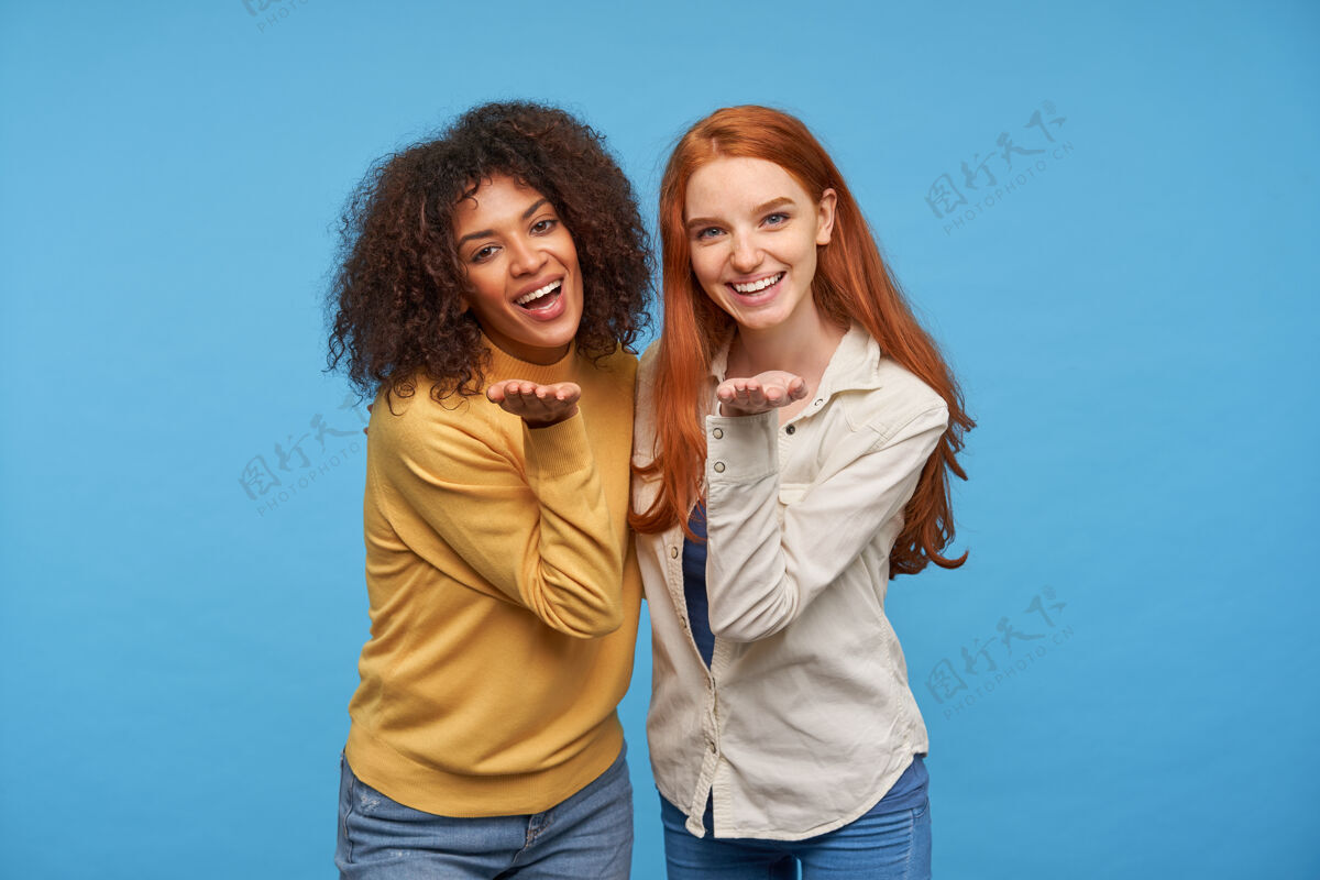 化妆品快乐迷人的年轻女朋友们互相拥抱 面带微笑 愉快地看着对方 在蓝色的墙壁上摆姿势时 掌心抬高自然女人室内