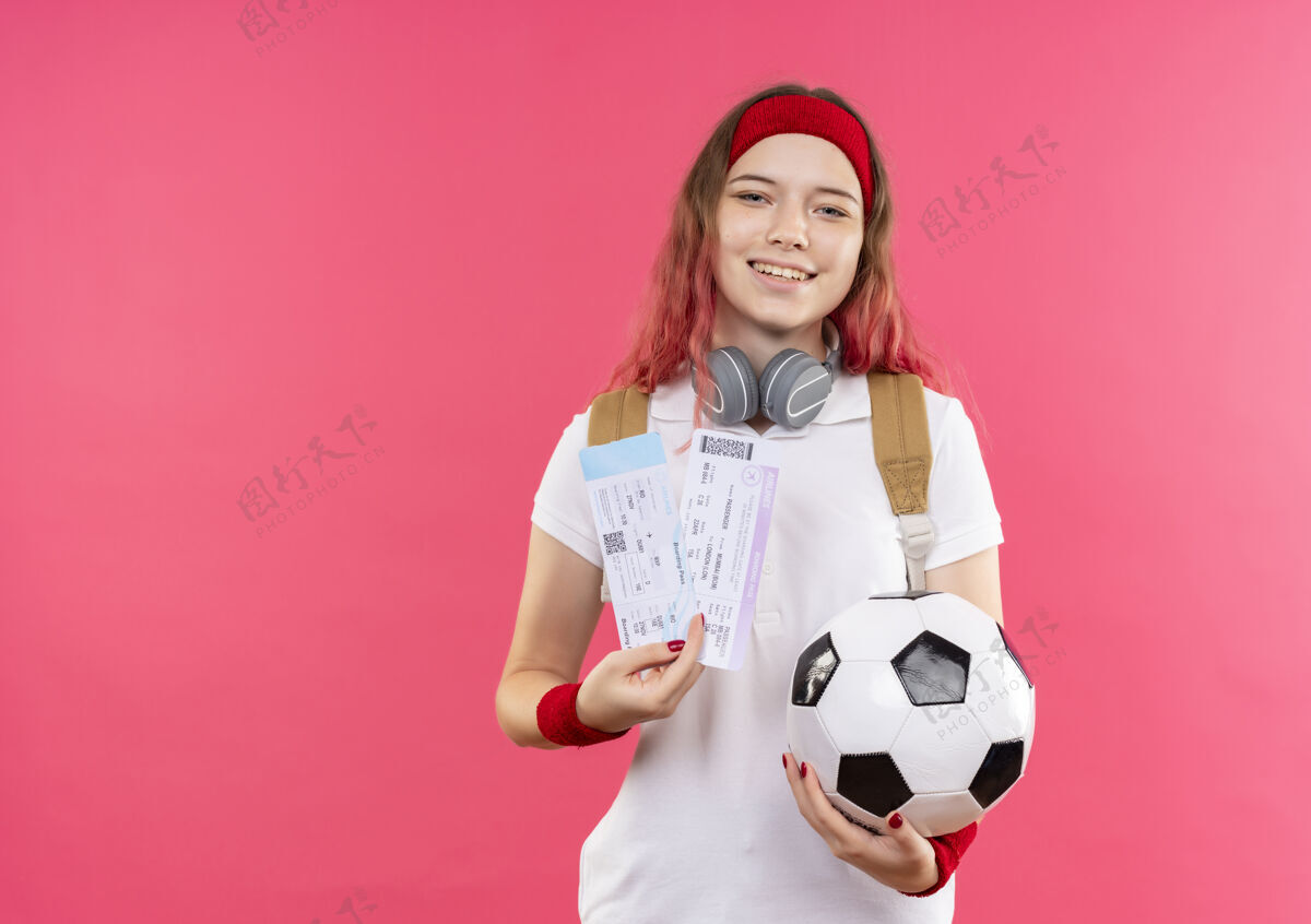 人戴着头巾 手持两张机票和一个足球的年轻运动女郎站在粉红色的墙上 脸上洋溢着愉快的笑容飞机微笑欢呼