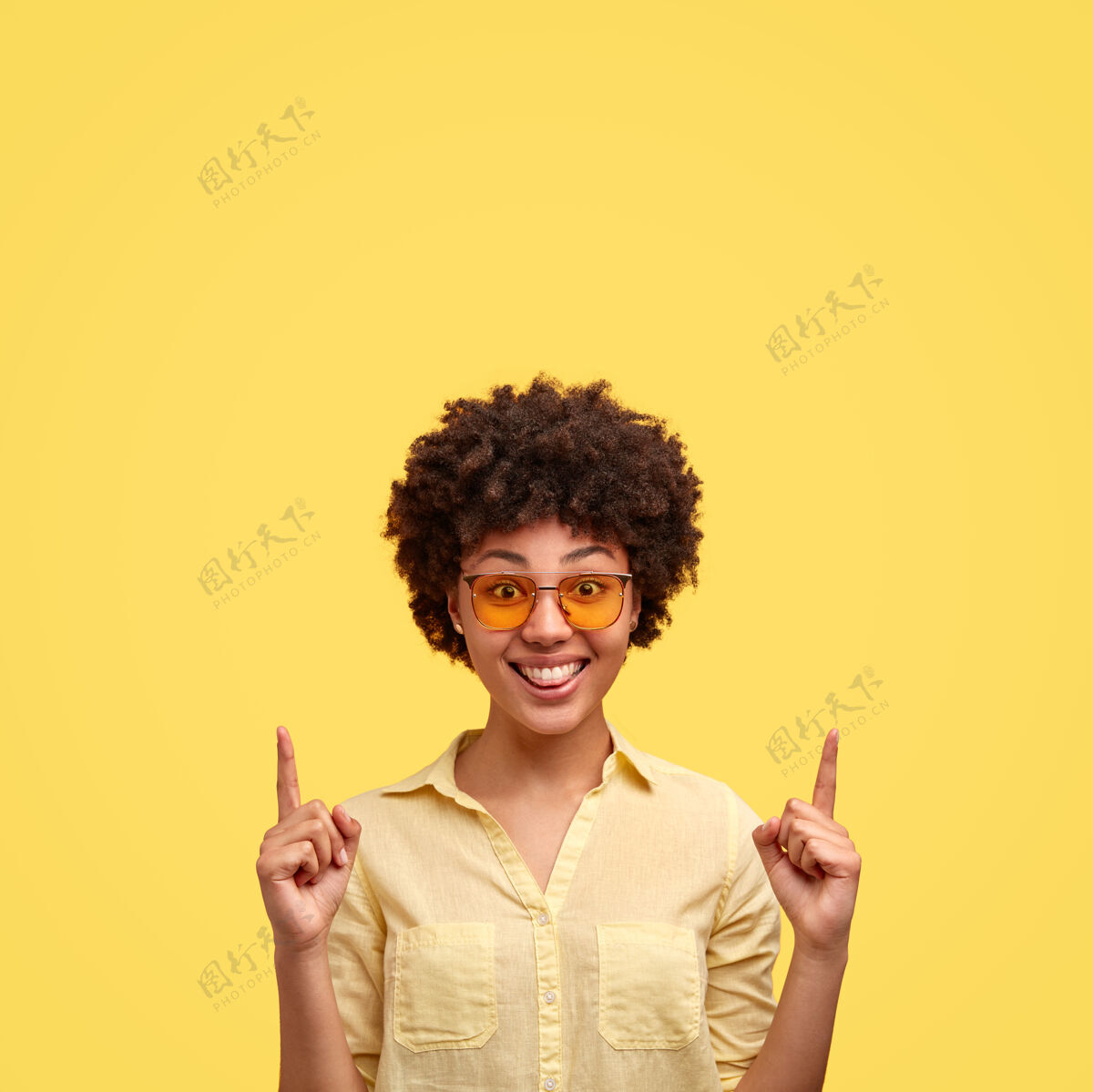 满意时尚黑人女性室内照有非洲式发型 穿着时髦的墨镜 衬衫与墙壁同色 正面微笑 向上表示头顶上方的自由空间用这个做广告肖像点高兴