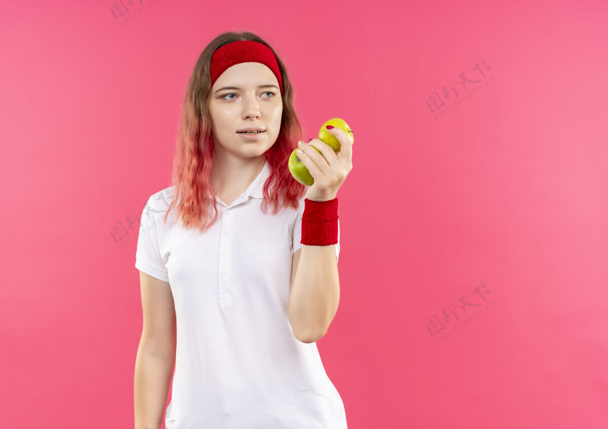 看戴着头巾 拿着两个苹果的年轻运动型女人站在粉红色的墙上 正快乐地看着一边运动站着人