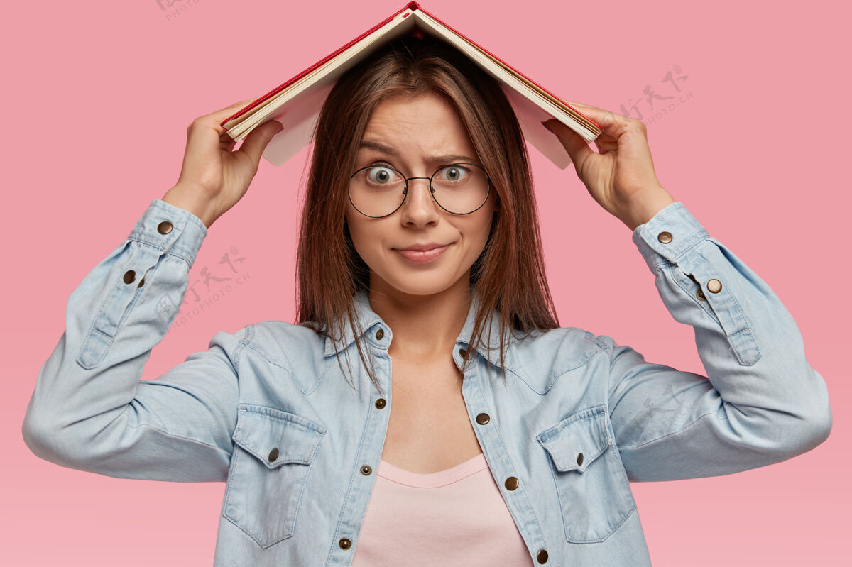 阅读不满的黑发女人表情不满 厌倦了不断的学习 把书放在头上 穿着牛仔衬衫 戴着眼镜 要求休假 隔着粉色的墙与世隔绝不满休闲文学