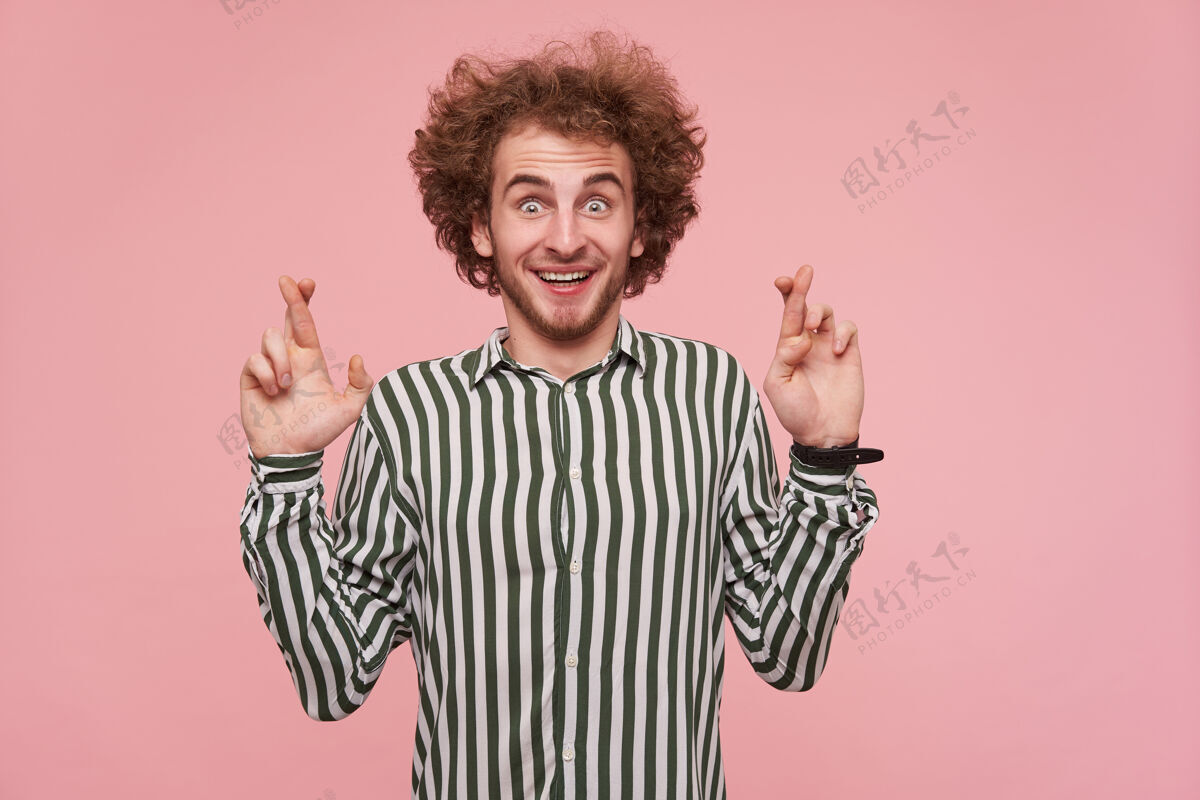 头发快乐的绿眼睛 卷曲的红头发的年轻男性 站在粉红色的墙上 开心地看着镜头 面带微笑 交叉着手指祈求好运发型提高未剃须