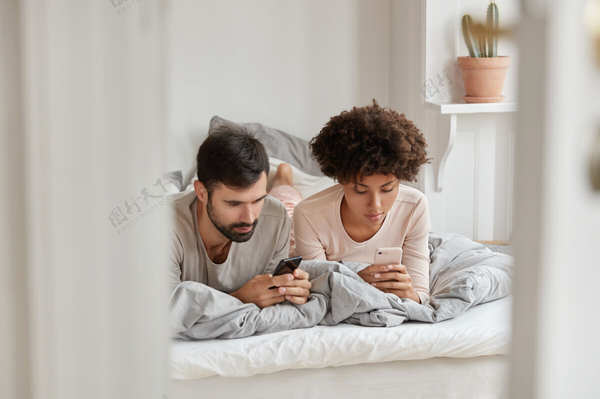 床男女混血的形象等待电话 躺在舒适的床上 在家休息 在社交网络上冲浪 在白天休息家庭 科技 休闲理念室内阅读成瘾