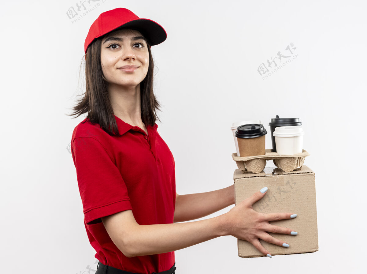 制服年轻的送货员穿着红色制服 戴着帽子 拿着盒子和咖啡杯 站在白色的背景下 面带微笑地看着相机拿着杯子年轻