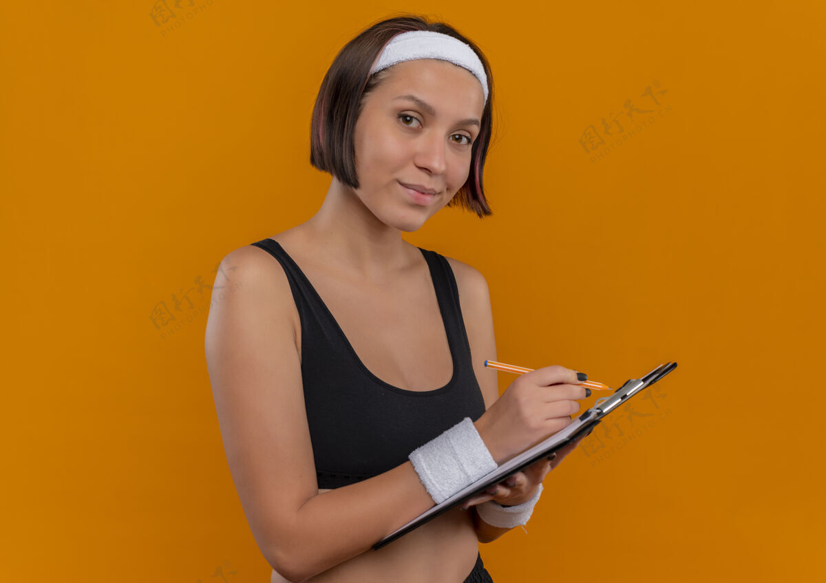 人身穿运动服的年轻健身女 头上戴着头巾 手里拿着写字板 空白的书页写着 站在橙色的墙上 看起来很自信公民运动员空白