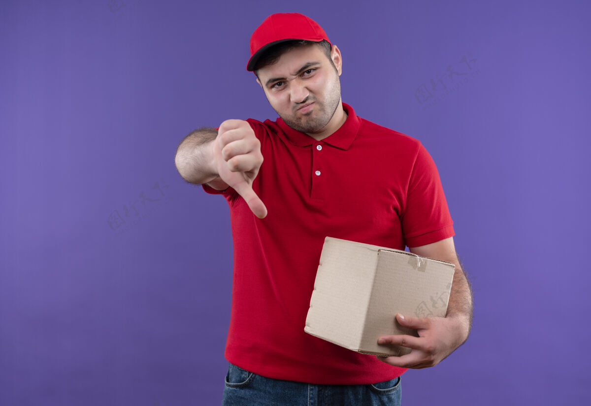 男人年轻的送货员穿着红色制服 戴着帽子 手里拿着一个盒子 站在紫色的墙上 大拇指朝下 皱着眉头脸皱眉公民