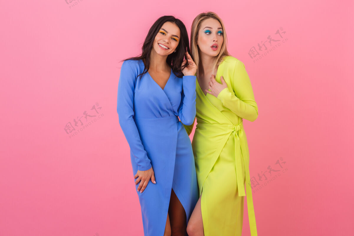 华丽两位时尚微笑迷人的女性朋友身着蓝黄相间的时尚多彩礼服 在粉色墙面上合影 成为春季时尚潮流折扣衣服多彩