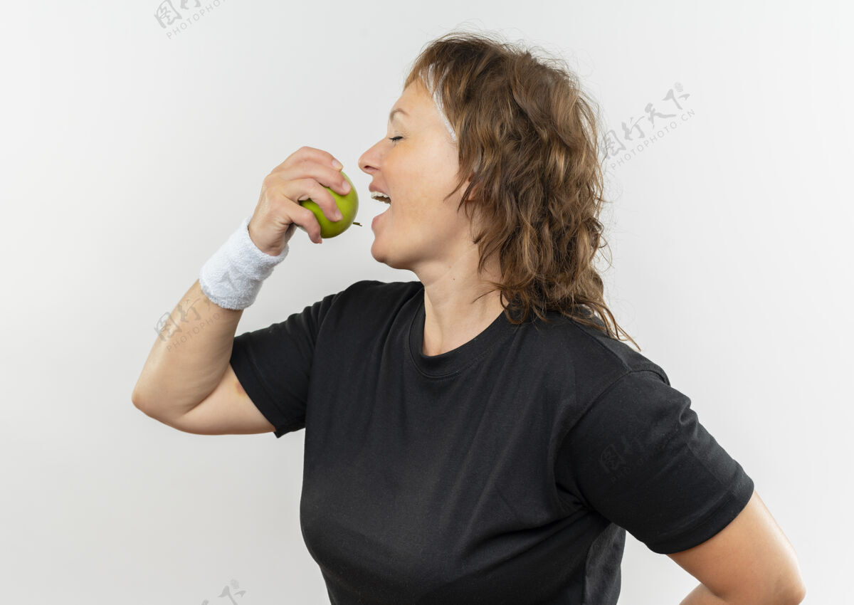站着中年运动女性 身穿黑色t恤 头上戴着绿色的苹果 咬着苹果 快乐的脸站在白色的墙上女人健康运动