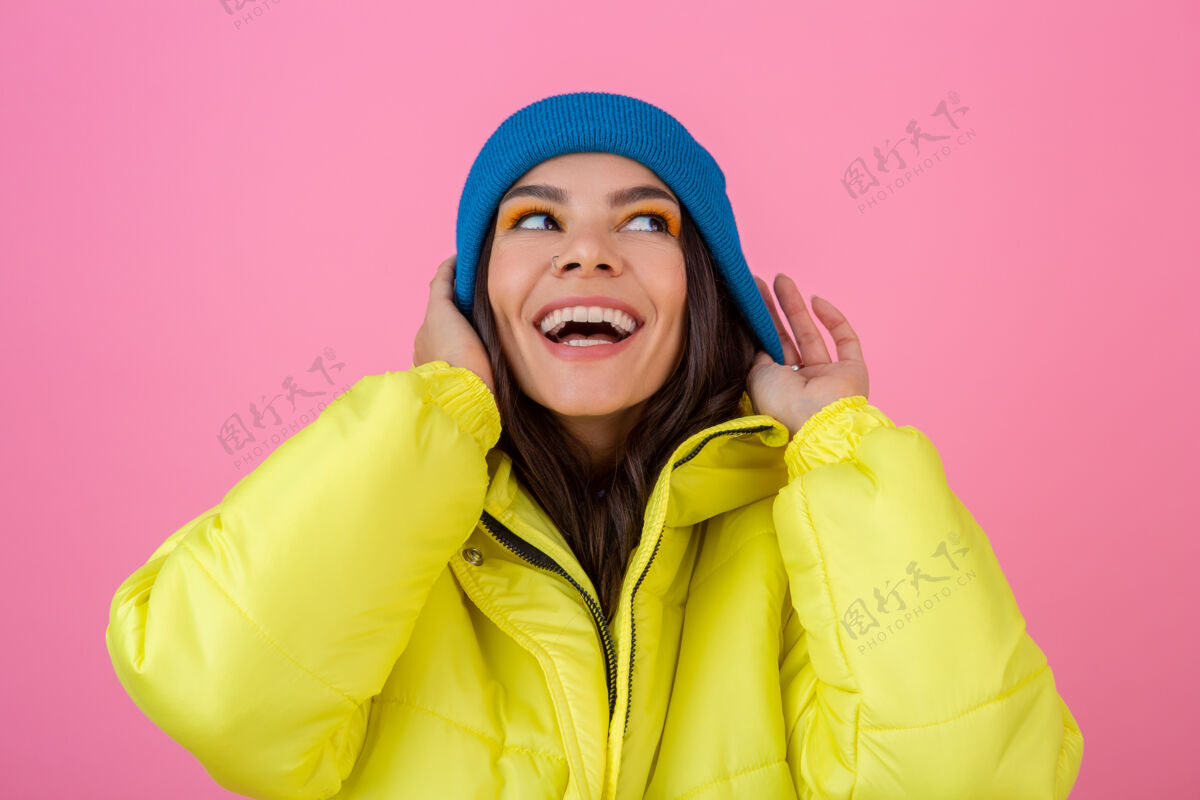 年轻一幅迷人微笑的时尚女人的画像 穿着黄色的彩色冬季羽绒服 戴着蓝色的针织帽子 穿着暖和的衣服 在粉色的墙上摆姿势 时尚潮流秋天夹克天气