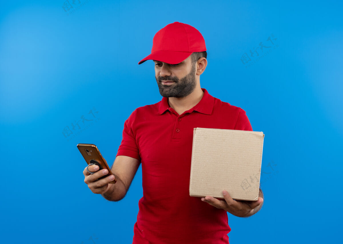 制服一个留着胡须 穿着红色制服 戴着帽子的送货员站在蓝色的墙上看着手机屏幕 严肃的脸胡子男人包装