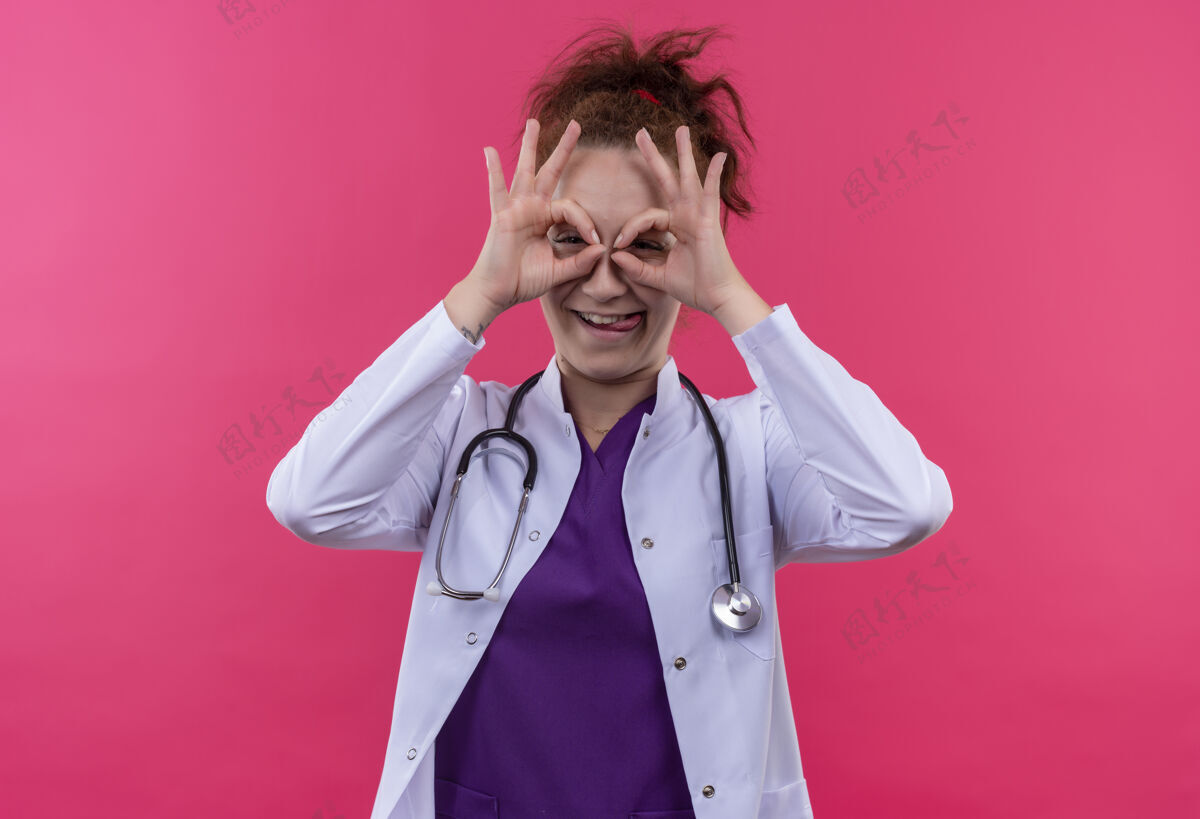 医生年轻的女医生 穿着白大褂 带听诊器 手指像望远镜一样 站在粉红色的墙上 伸出舌头 透过手指看 做着正常的手势女人穿过望远镜