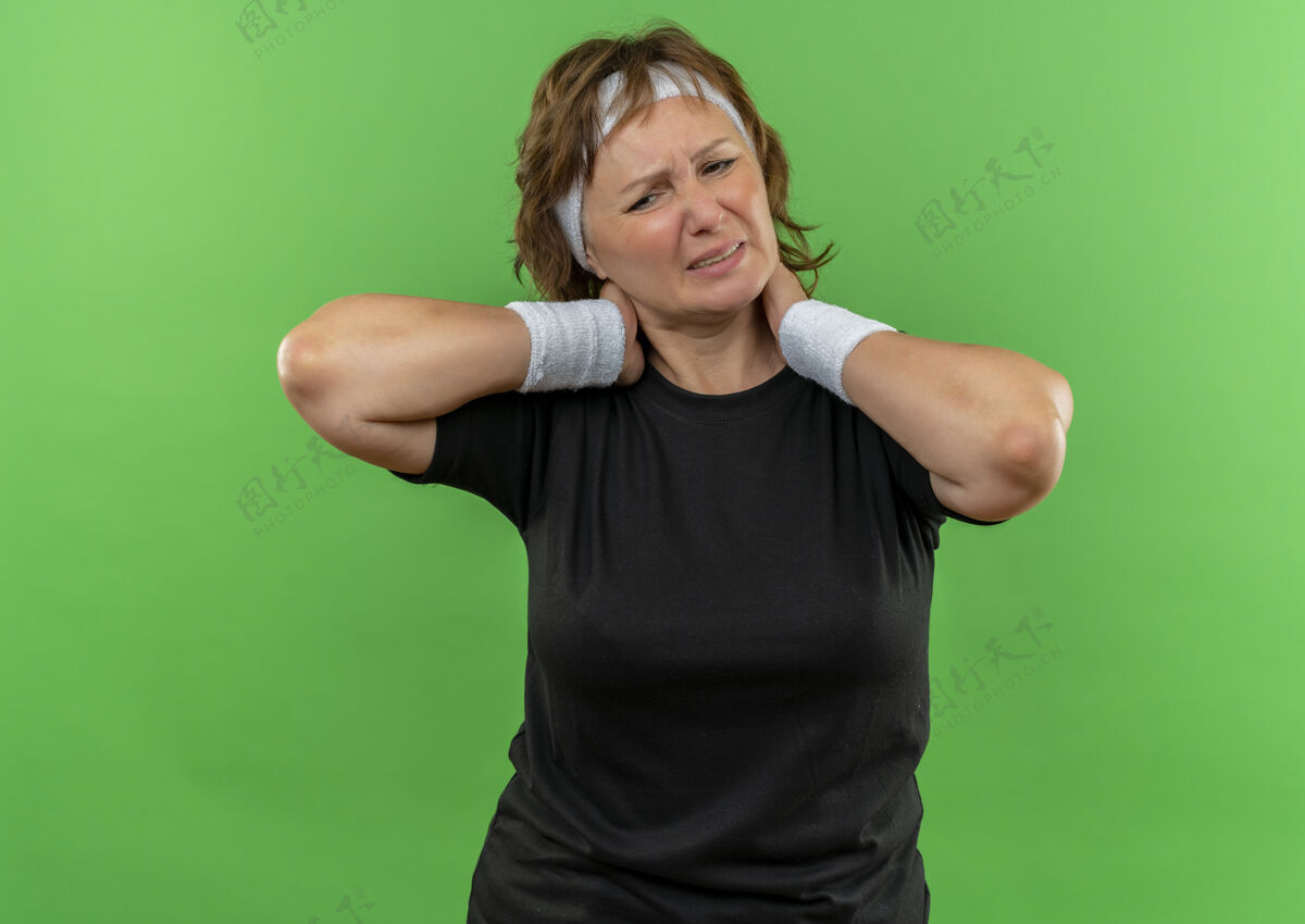 运动员中年运动女性 身穿黑色t恤 头带看起来不舒服 摸着脖子 站在绿色墙壁上感觉疼痛市民老年人中年人