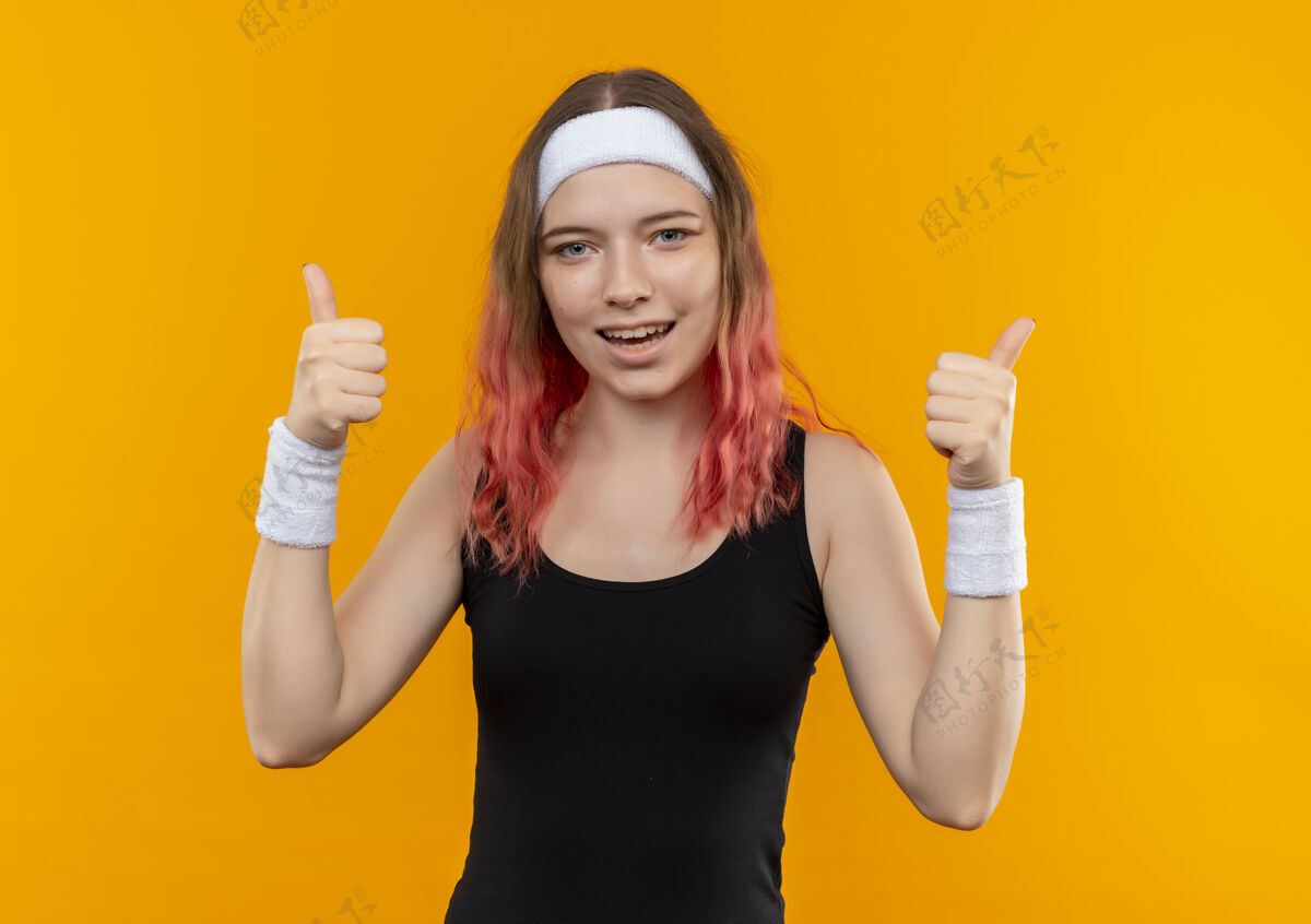 女人身着运动服的年轻健身女士微笑着 双手竖起大拇指站在橙色的墙上人拇指手