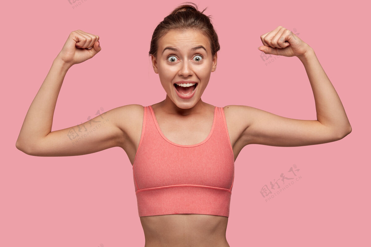皱眉图为强壮开朗的年轻女运动员在健身房锻炼肌肉 穿着与墙壁同色调的休闲上衣 拥有健康完美的身体 令人惊叹运动理念衣服时尚二头肌