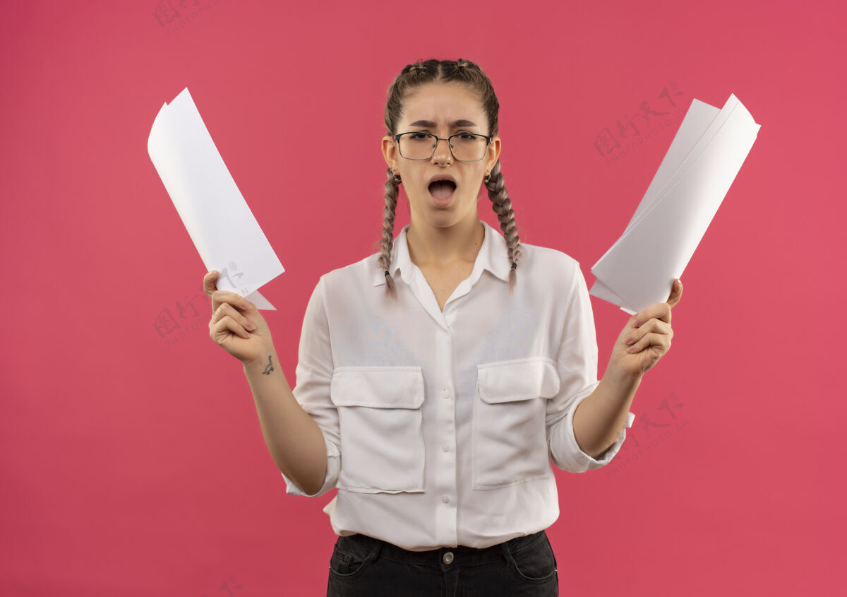 眼镜戴着眼镜 梳着辫子 穿着白衬衫的年轻女学生拿着空白页望着前面 失望地举起双臂站在粉色的墙上站着学生摆姿势