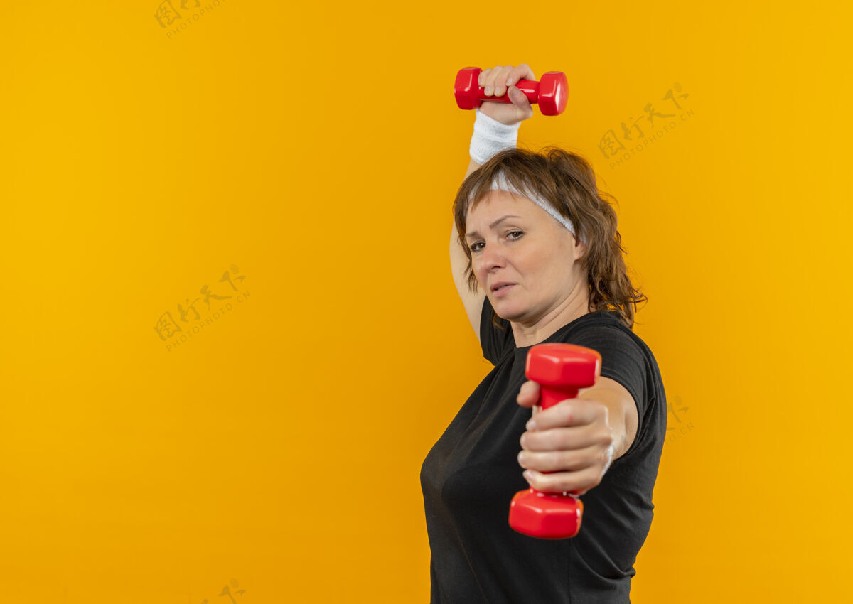 长相身穿黑色t恤 头箍和哑铃的中年运动女性站在橙色的墙上 显得自信而紧张年龄中年工作