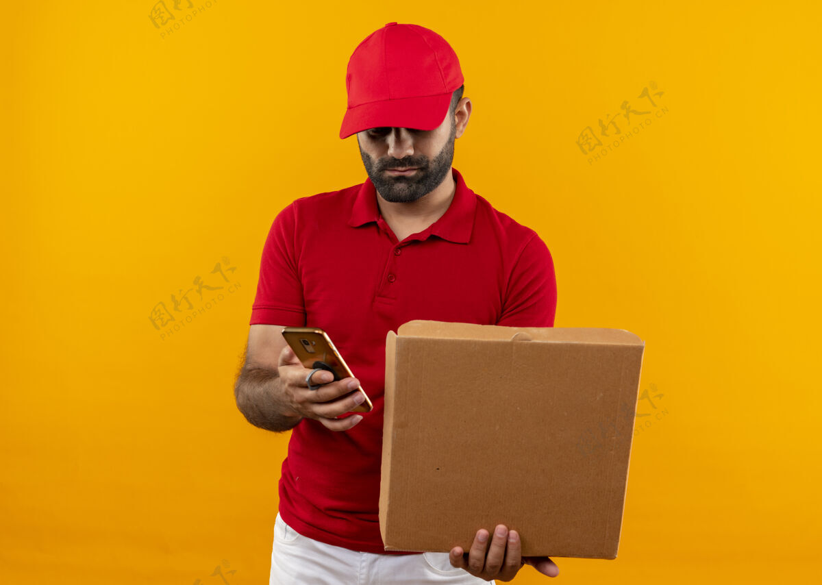 帽子留着胡须的送货员身穿红色制服 戴着帽子 手里拿着打开的比萨饼盒 站在橙色的墙上用手机发短信拿着电话男人