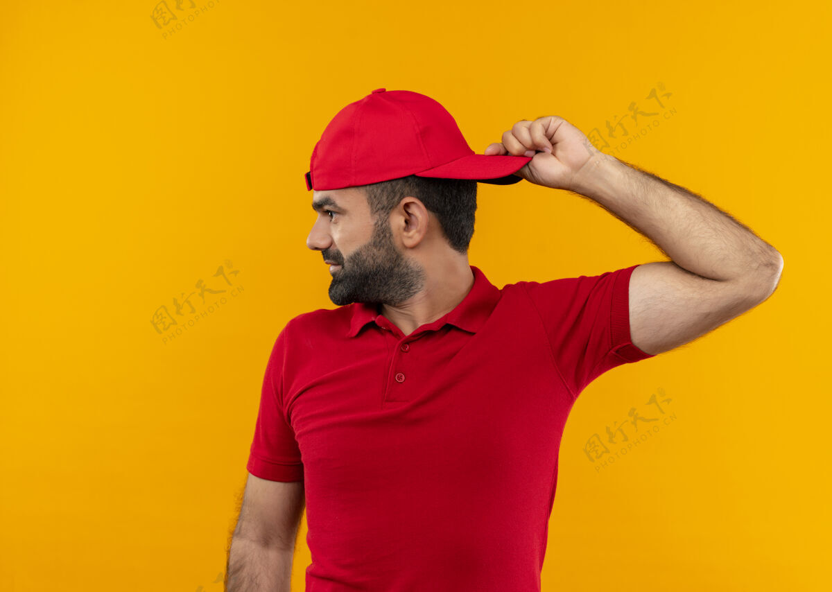 微笑一个留着胡须 身穿红色制服 戴着帽子的送货员微笑着把帽子翻过来 站在橙色的墙上男人靠边转身