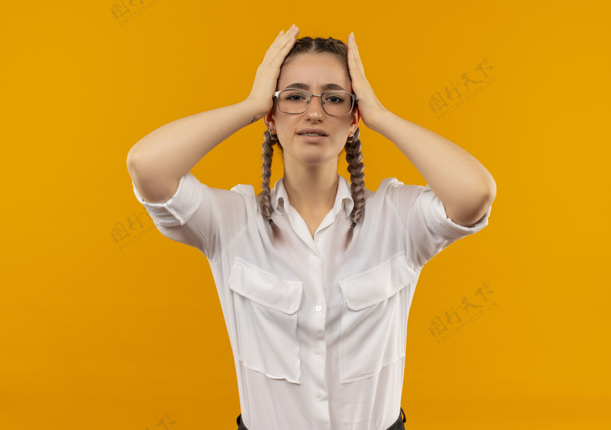 头一个戴着眼镜 梳着辫子 穿着白衬衫的年轻女学生站在橙色的墙上 抱着头 看上去很困惑眼镜姿势困惑