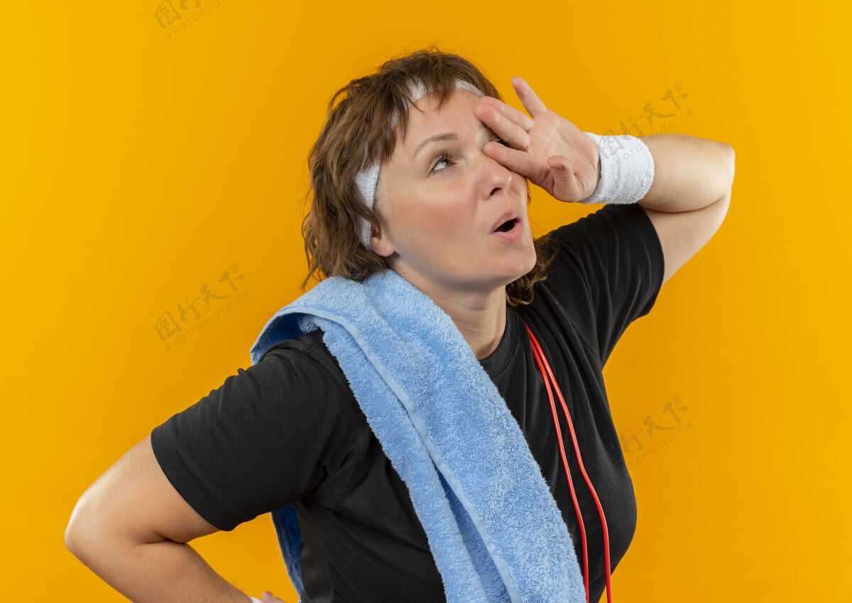 疲倦中年运动女性 身穿黑色t恤 头上系着头带 肩上搭着毛巾 站在橙色的墙上锻炼后 看上去既疲惫又疲惫肩膀运动老年