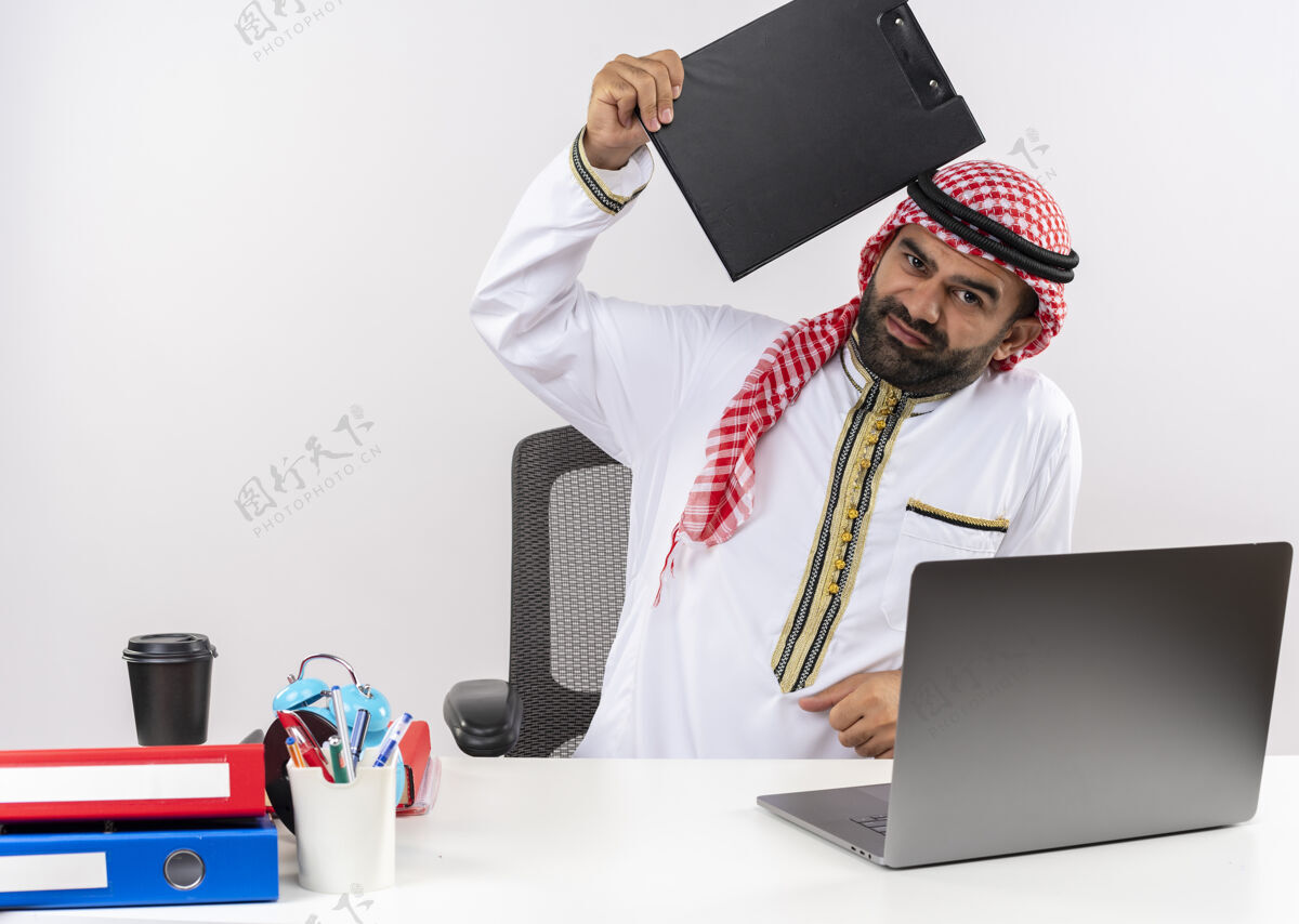 剪贴板不高兴的阿拉伯商人穿着传统服装坐在桌旁 手提电脑拿着剪贴板 带着负面情绪在办公室工作情绪工作传统