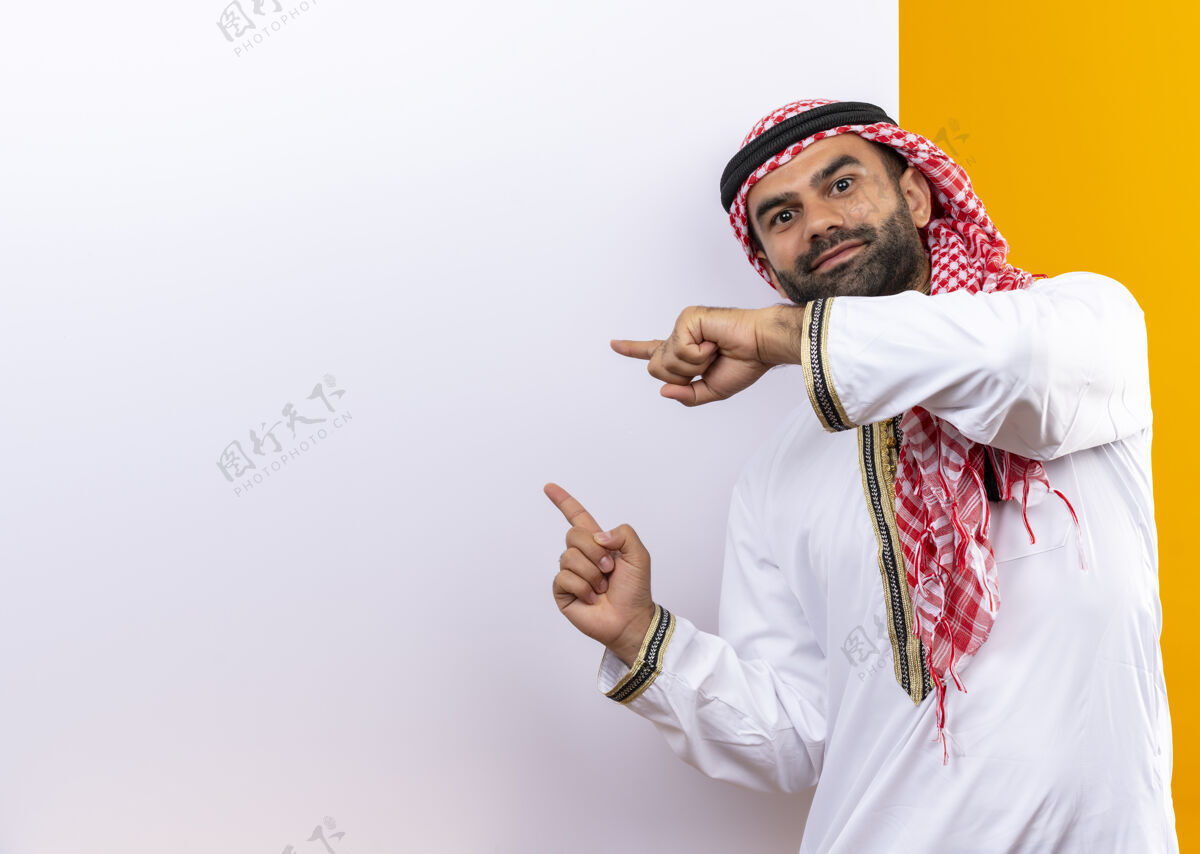 传统身着传统服装的阿拉伯商人站在空白广告牌旁 手指指着广告牌 脸上带着自信的微笑 越过橙色的墙壁脸指点商务人士