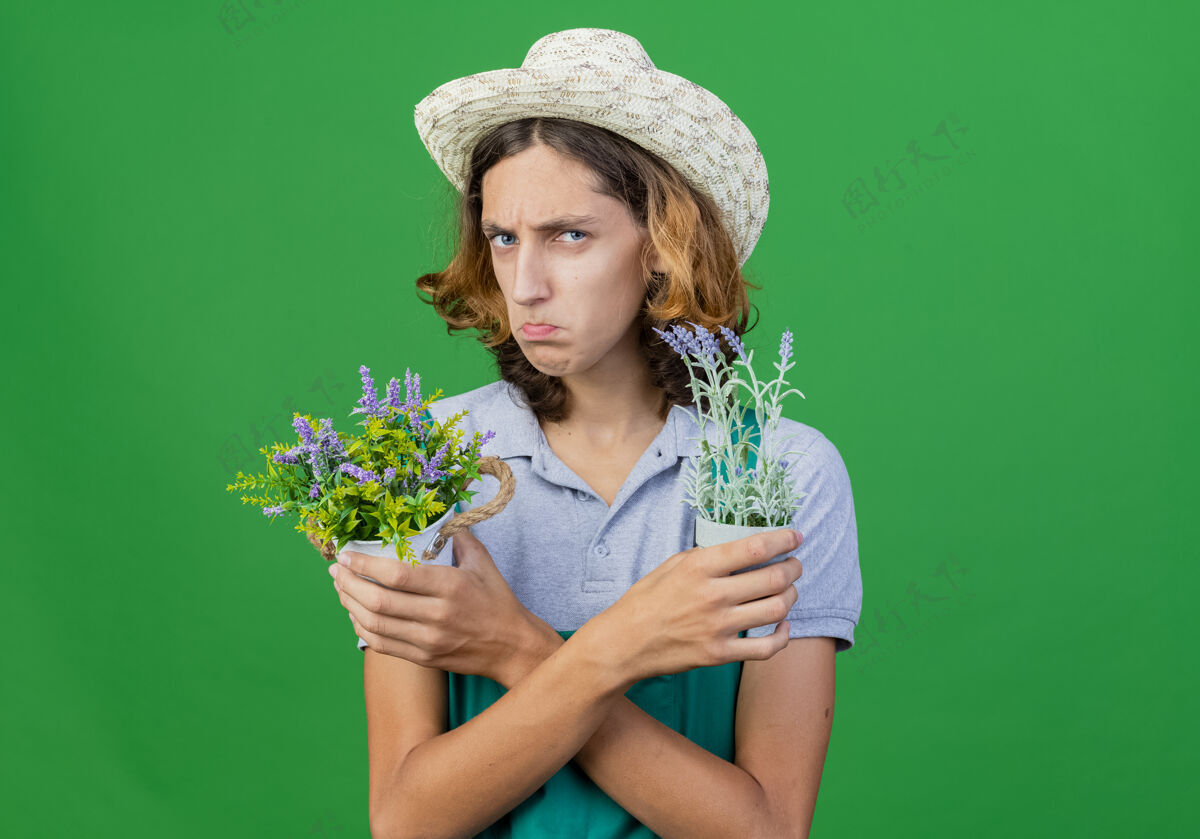 脸年轻的园丁 穿着连体衣 戴着帽子 手里拿着盆栽植物穿着抱着植物