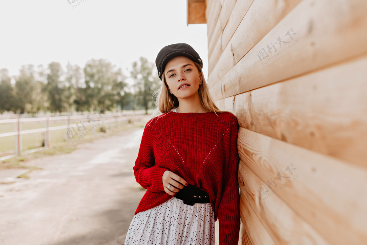 毛衣穿着红毛衣戴着黑帽子的金发美女在木墙附近的公园里摆姿势秋天穿着时令流行服装的美女时尚外套年轻