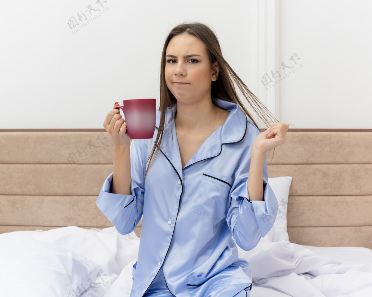 杯子穿着蓝色睡衣的年轻漂亮女人坐在床上 端着一杯咖啡 看着摄像机 不高兴地在卧室里的灯光背景下醒来年轻蓝色漂亮