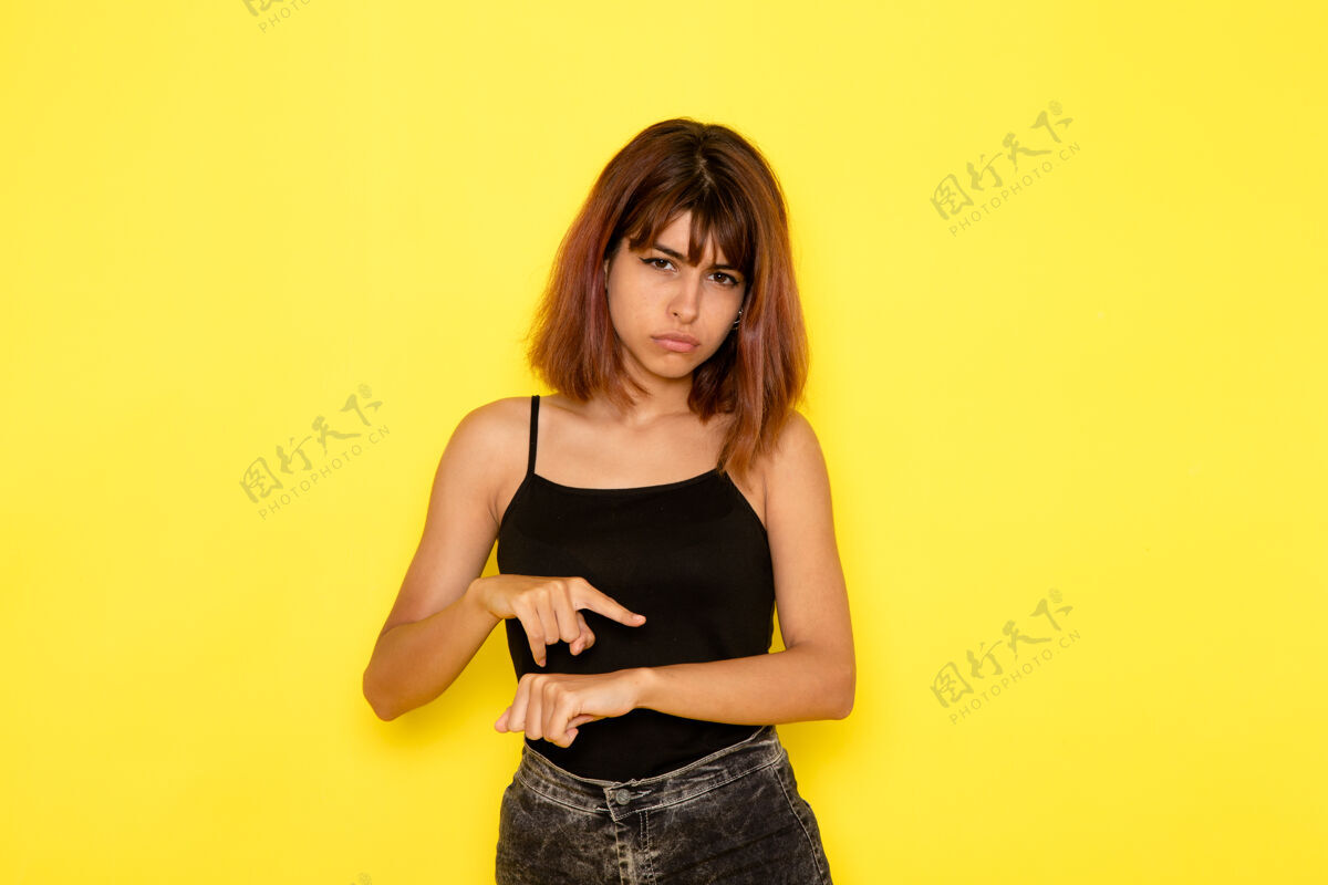 年轻身穿黑色衬衫和灰色牛仔裤的年轻女性在黄色墙上展示手腕的正面视图姿势漂亮模特
