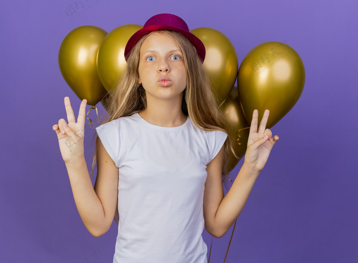 看戴着节日礼帽的漂亮小女孩和一群巴隆人看着镜头 开心而积极地展示着v型标志 紫色背景下站着生日派对的概念束气球节日
