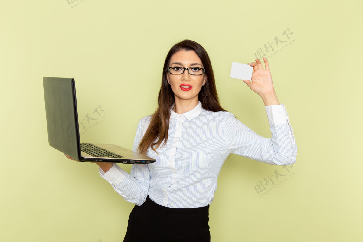 微笑穿着白衬衫和黑裙子的女上班族在浅绿色墙上使用笔记本电脑和名片的前视图女前面拿着