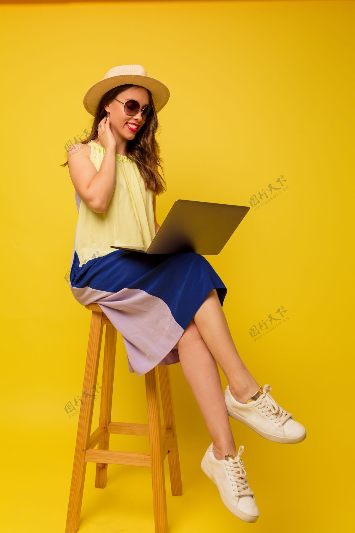 嘴唇积极时尚的女人戴着帽子 穿着夏装 在黄色的墙上用笔记本电脑工作学生乐观微笑