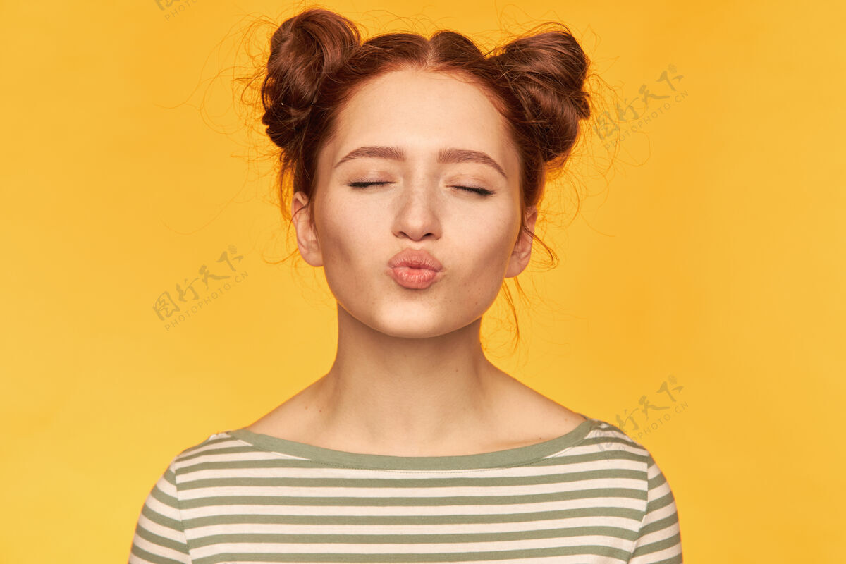 年轻迷人可爱的红发女孩的肖像 两个发髻 健康的皮肤闭上眼睛的甜蜜亲吻穿着条纹毛衣 孤立地站着 在黄色的墙上特写女人时尚皮肤