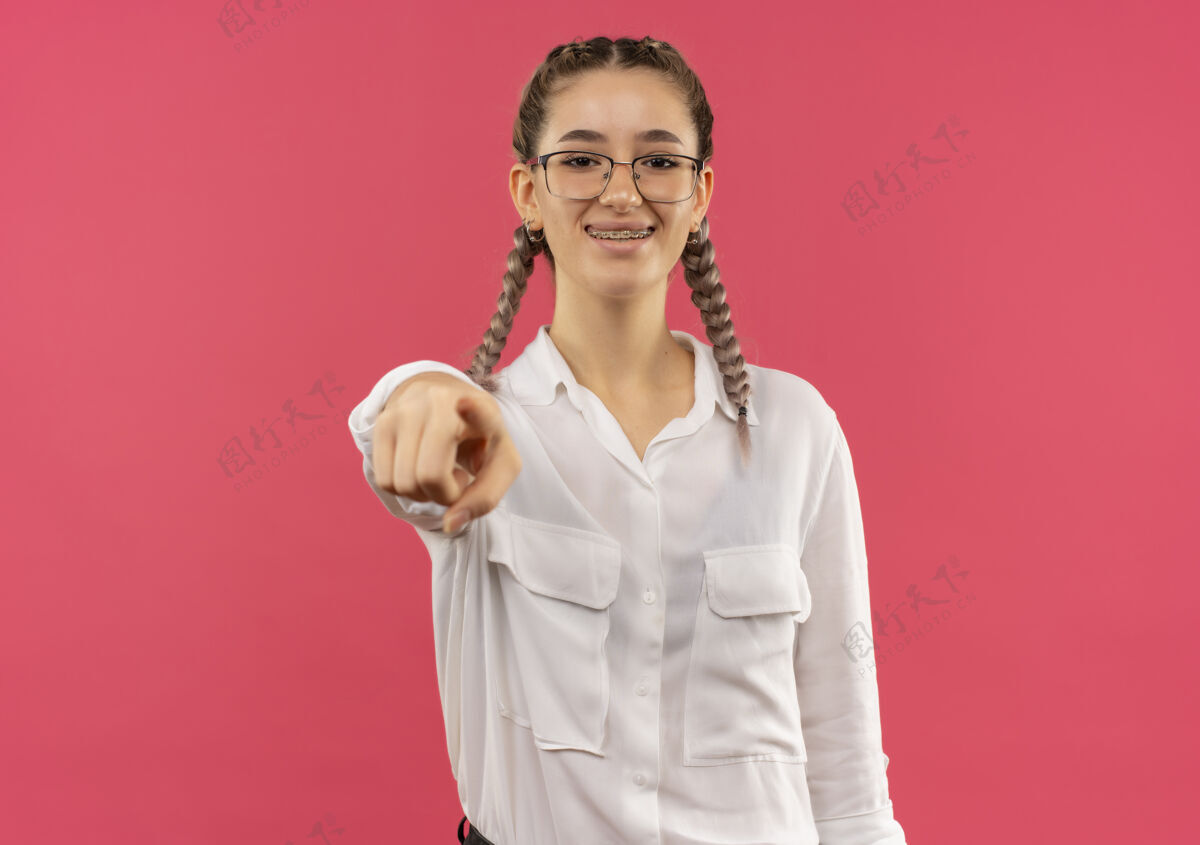 眼镜戴着眼镜 梳着白衬衫辫子的年轻女学生用食指指着前面 微笑着自信地站在粉色的墙上衬衫学生手指