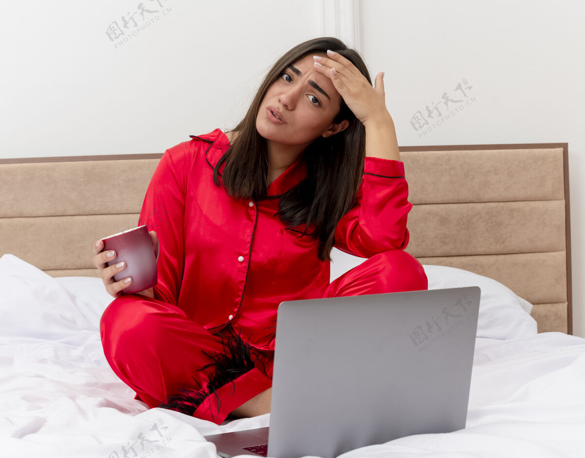 不高兴穿着红色睡衣的年轻漂亮女人坐在床上 拿着笔记本电脑和一杯咖啡 看着摄像机 在卧室内部的灯光背景下感到困惑和不快室内看着睡衣