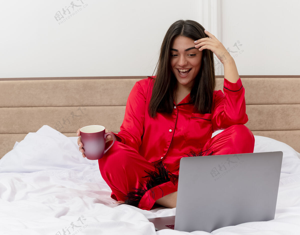 咖啡穿着红色睡衣的年轻美女坐在床上 手里拿着笔记本电脑和一杯咖啡 在卧室的室内灯光背景下愉快地微笑着杯子女士红色