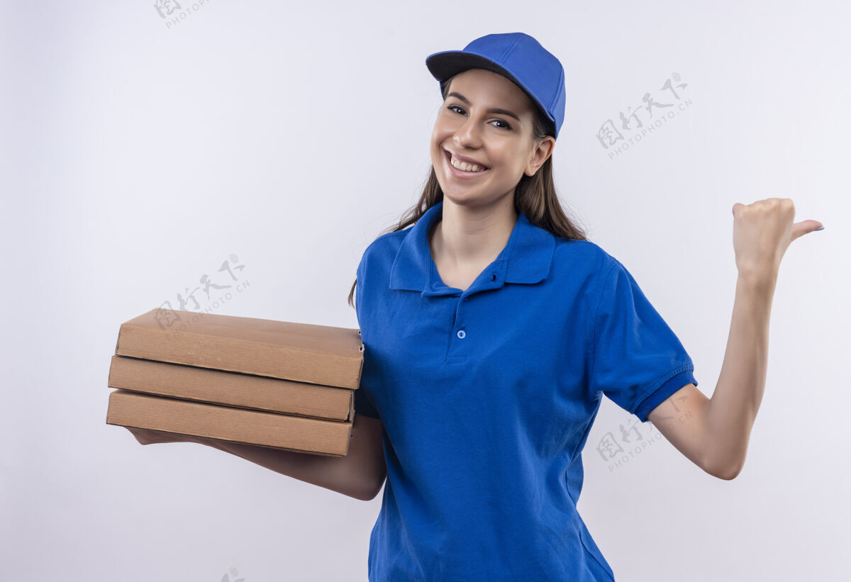 披萨穿着蓝色制服 戴着帽子的年轻送货女孩拿着一叠披萨盒 拇指朝后 愉快地微笑着年轻制服拇指
