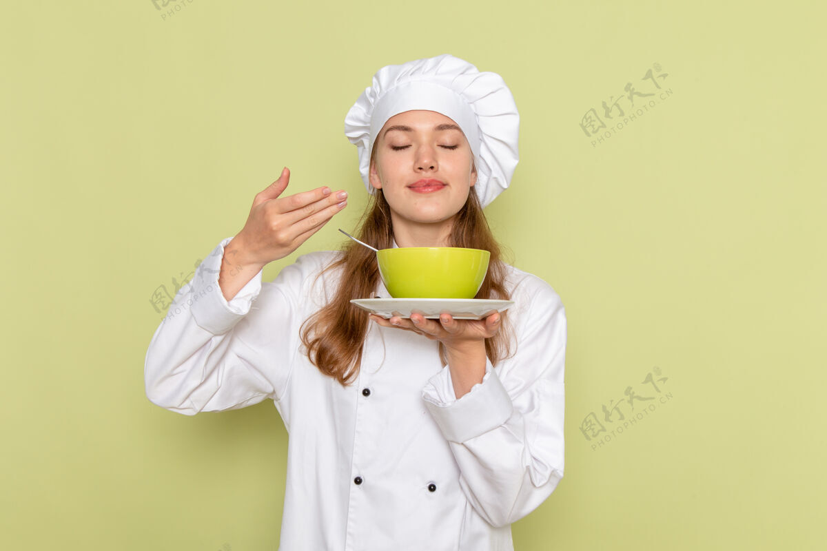 前面身穿白色厨师服的女厨师手持绿色盘子 在绿色墙上闻着味道女帽子食物