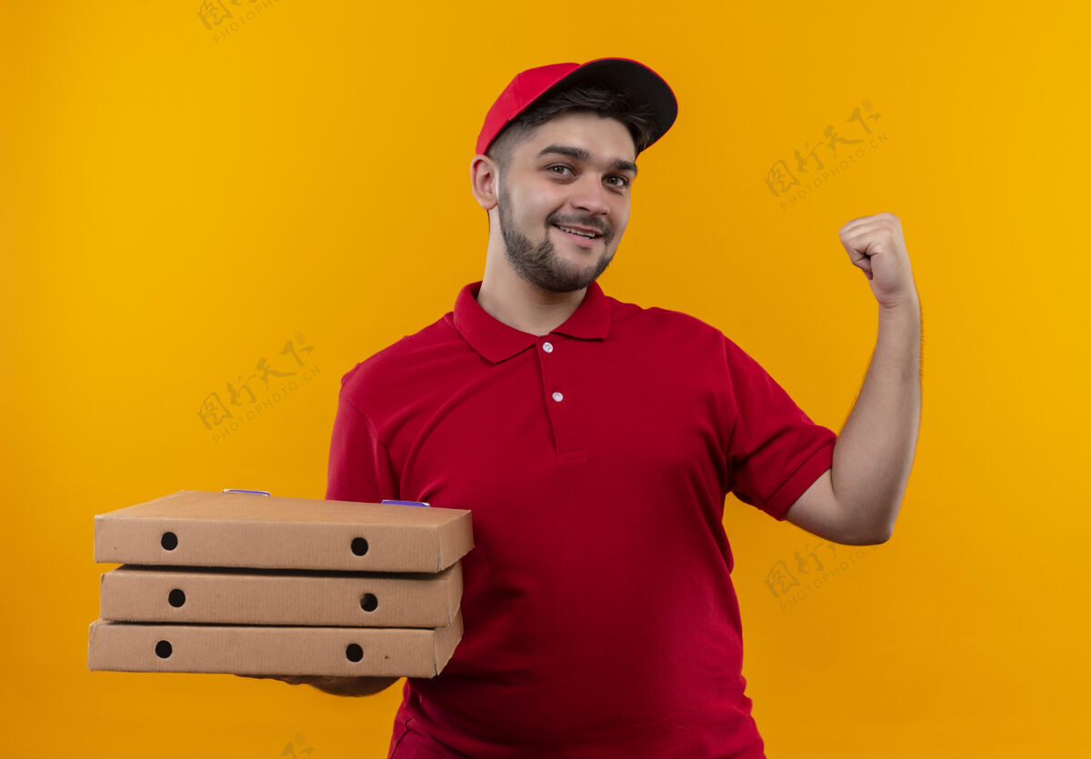自信年轻的送货员穿着红色制服 戴着帽子 手里拿着一叠比萨饼盒 自信地微笑着指着后面年轻人披萨背