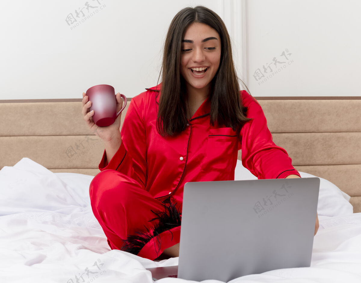 坐着穿着红色睡衣的年轻美女坐在床上 手里拿着笔记本电脑和一杯咖啡 在卧室的室内灯光背景下愉快地微笑着年轻红色快乐