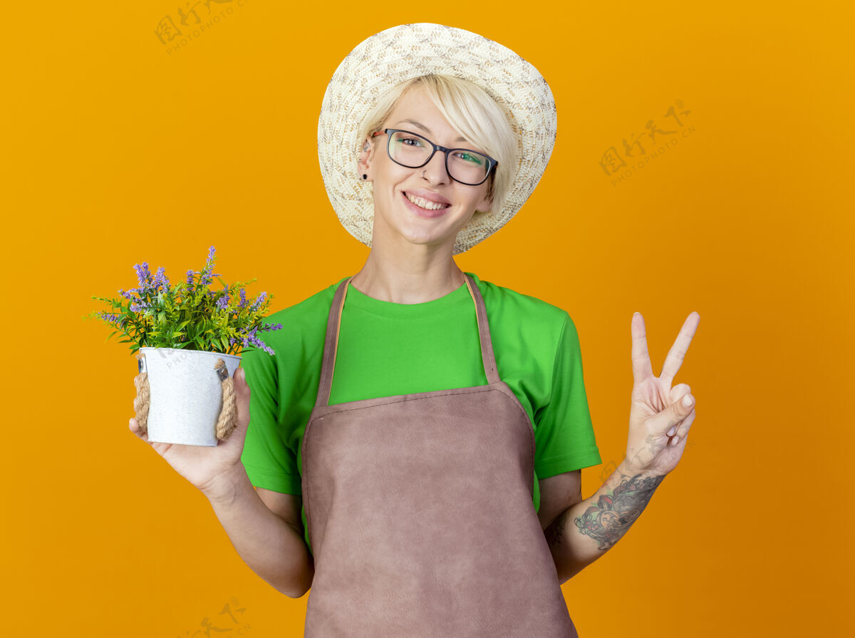 相机一位年轻的园丁 围裙上留着短发 戴着帽子 手里拿着盆栽植物 面带微笑地看着摄像机 橙色背景上有一个v形标志短裤帽子锅