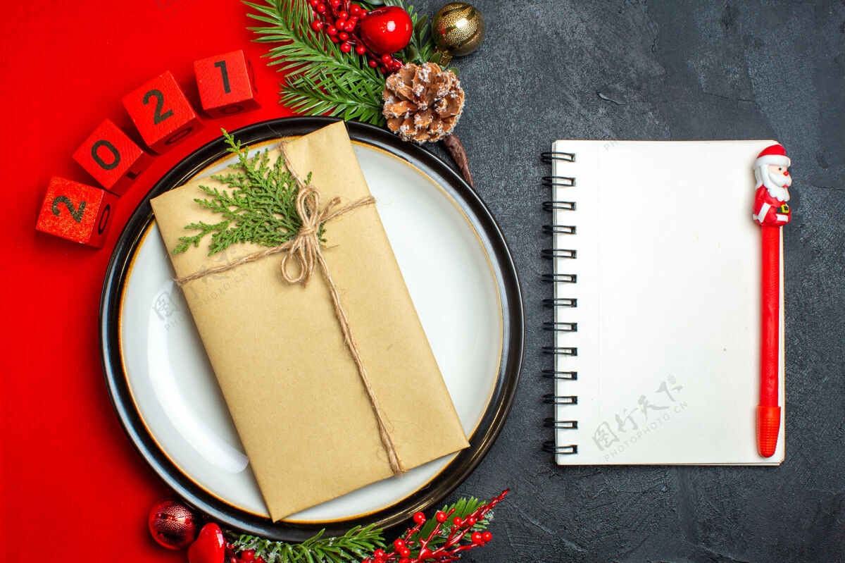 笔记本新年背景俯视图 餐盘上有礼物装饰配件杉木树枝和数字 红色餐巾上有笔记本 黑色桌子上有钢笔餐盘数字圣诞