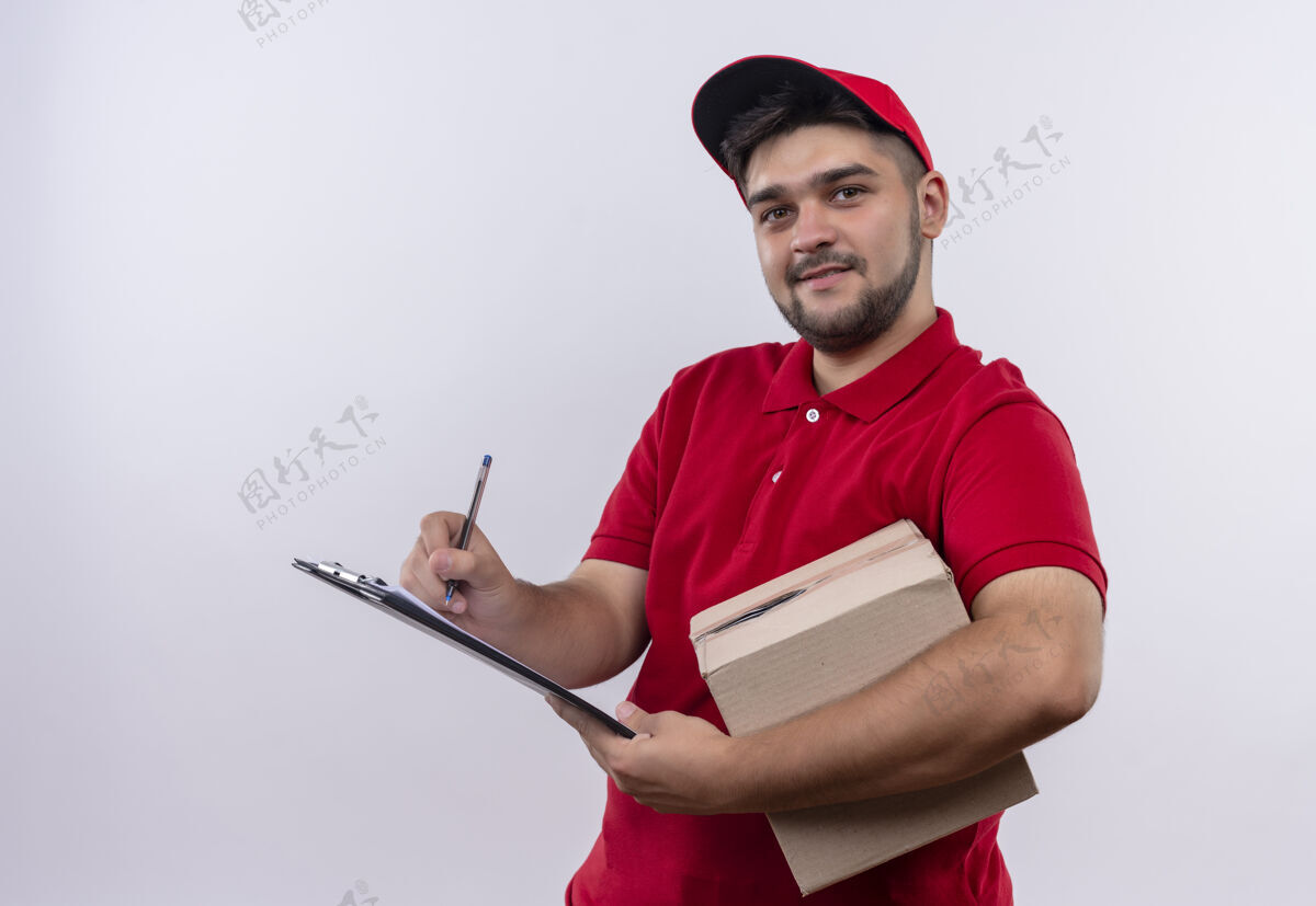 朋友年轻的送货员穿着红色制服 戴着帽子 拿着盒子 在空白页上写着友好的微笑制服红色年轻