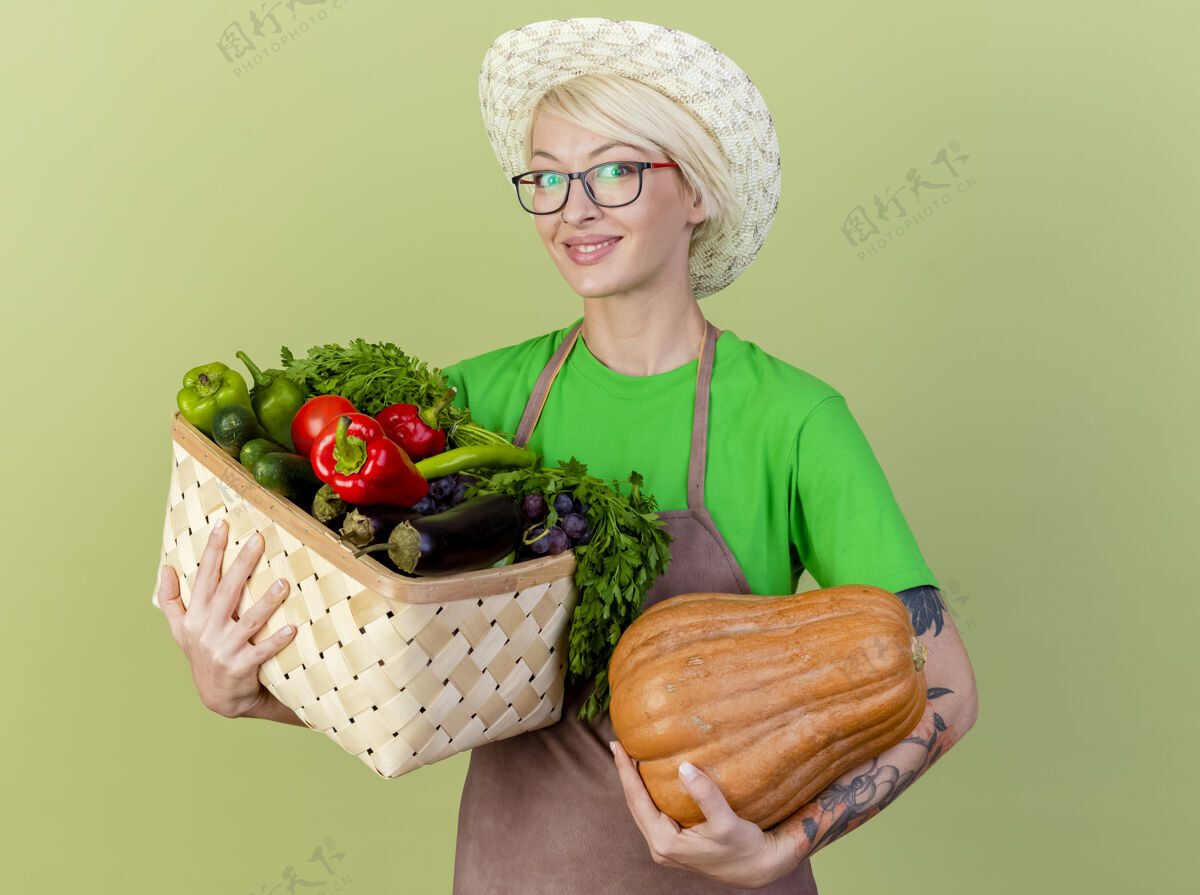 女人围裙里留着短发 戴着帽子 手里拿着南瓜和装满蔬菜的箱子 看着摄像机 站在明亮的背景下开心地微笑着抱围裙充分