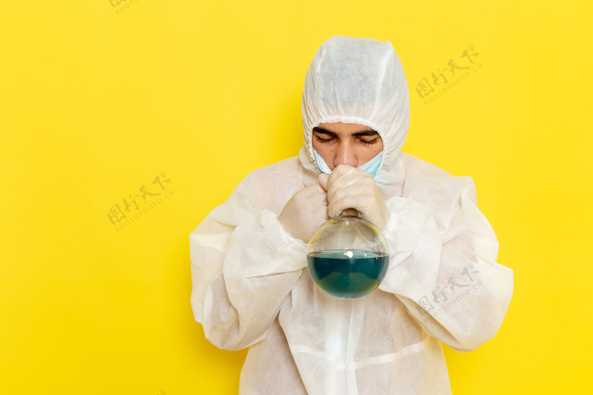 科学穿着特殊防护服的男科学工作者的正面图 拿着装有溶液的烧瓶 在黄色的墙上闻着男性溶液理科