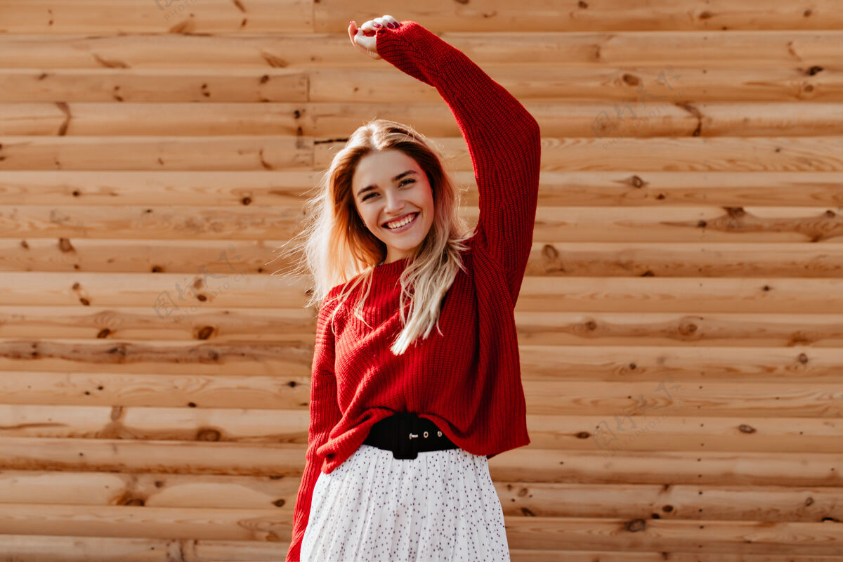 金发漂亮的金发女郎穿着漂亮的红色毛衣在户外玩得很开心微笑的年轻女子在木墙上快乐地摆着姿势肖像身体女人