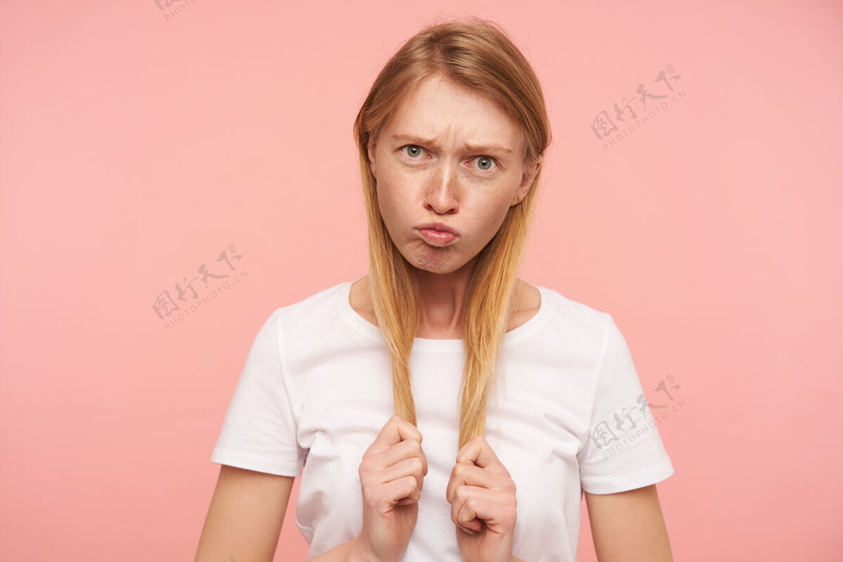 傻瓜有趣的镜头 年轻的绿眼长发红发女性穿着白色的基本t恤 噘着嘴唇和鬼脸 同时愚弄粉红色的背景室内情绪长