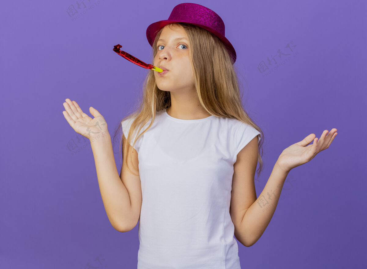 看美丽的小女孩在节日帽子吹哨子看起来困惑 生日聚会的概念站在紫色的背景节日漂亮困惑