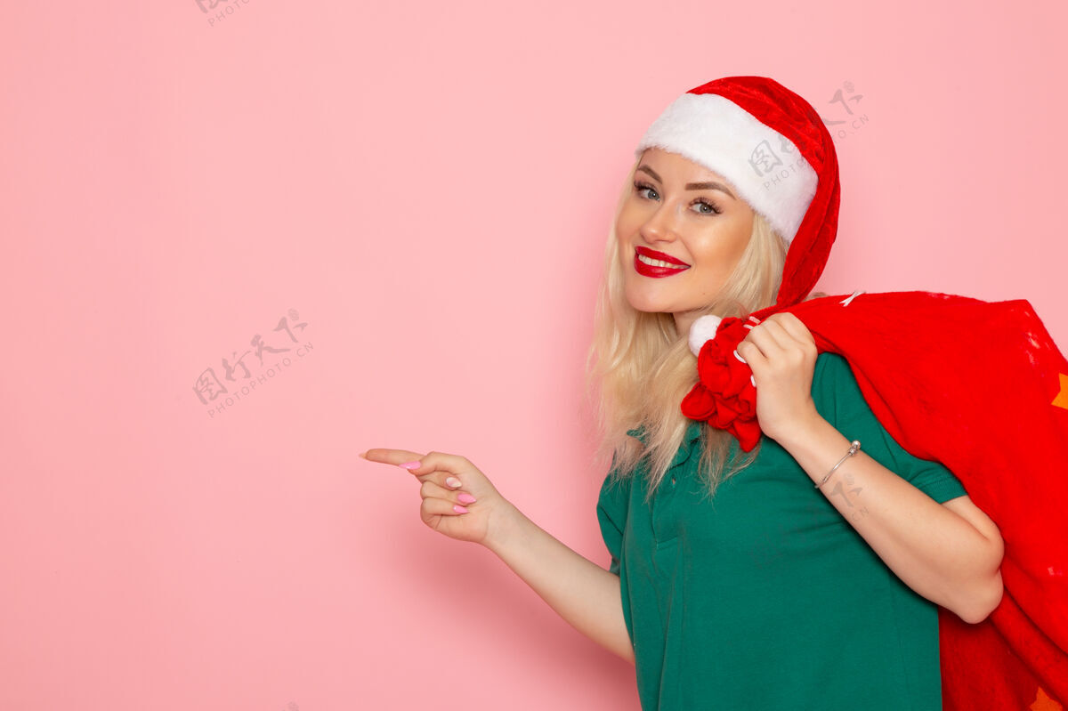 年轻的女性正面图年轻女性提着红包带着礼物在粉色墙上度假模型圣诞老人新年照片携带金发肖像